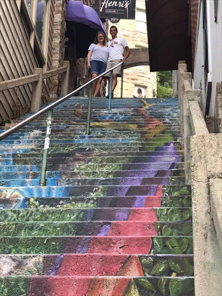 A colorful mural decorates a stairway where a couple poses beside a handrail.