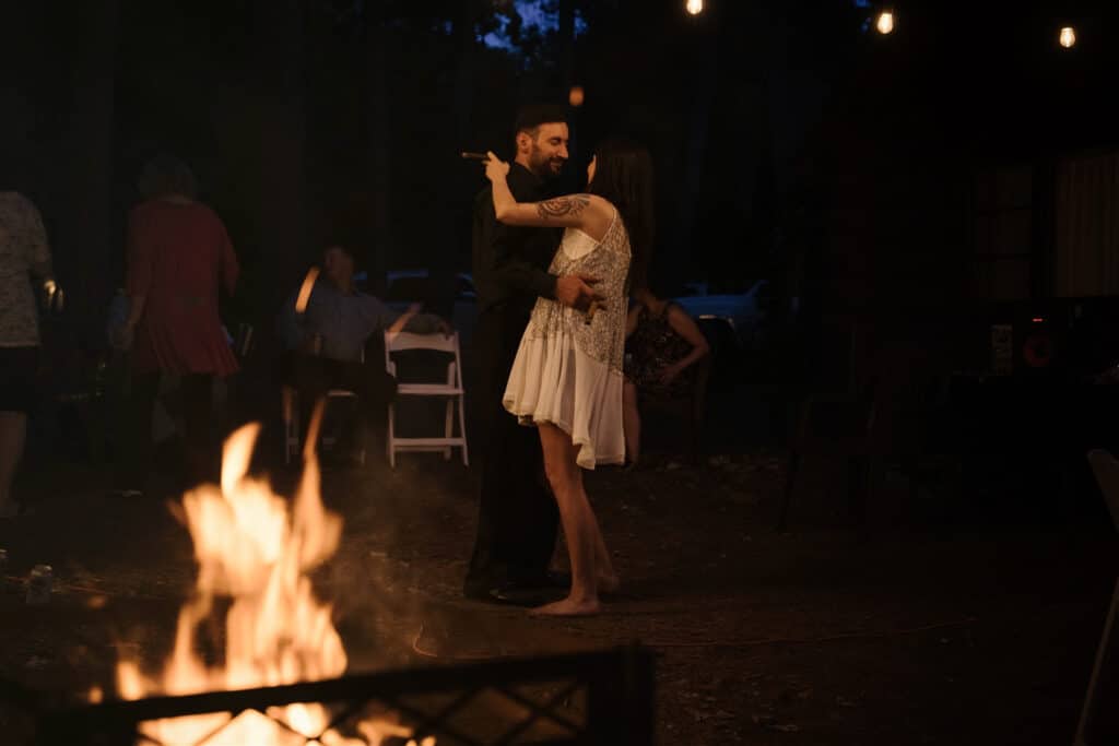 A couple dances together by a fire at dusk, with others in the background.