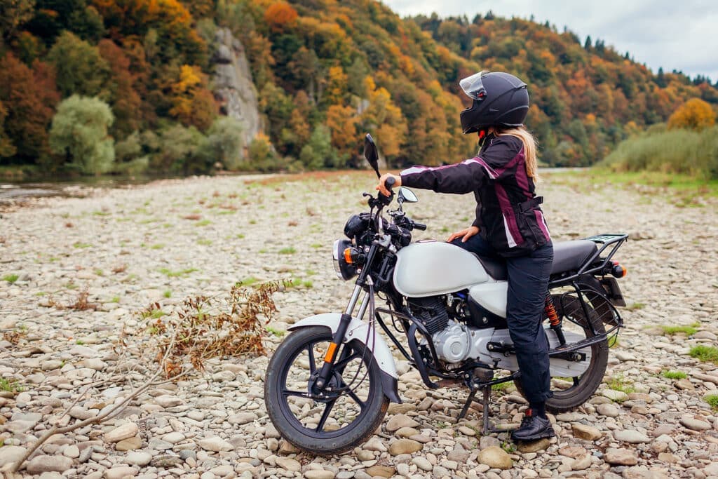 A motorcyclist in a helmet stands beside a white motorcycle on a rocky riverbank surrounded by colorful autumn foliage.