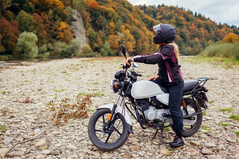 A motorcyclist in a helmet stands beside a white motorcycle on a rocky riverbank surrounded by colorful autumn foliage.