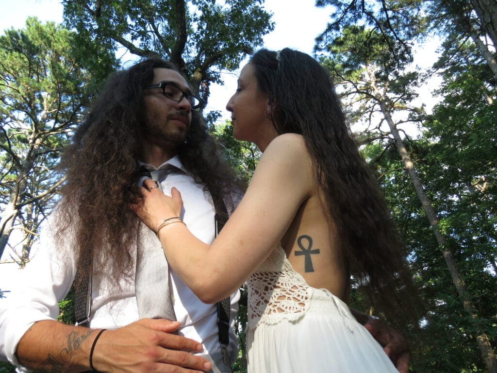 A couple shares an intimate moment in a wooded setting, with the woman wearing a backless white dress and the man in a suit and tie.