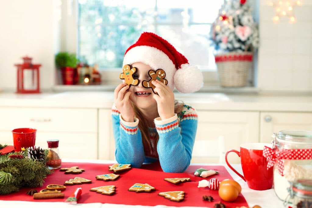 A child in a Santa hat playfully holds gingerbread cookies to her eyes while smiling at a festive table.
