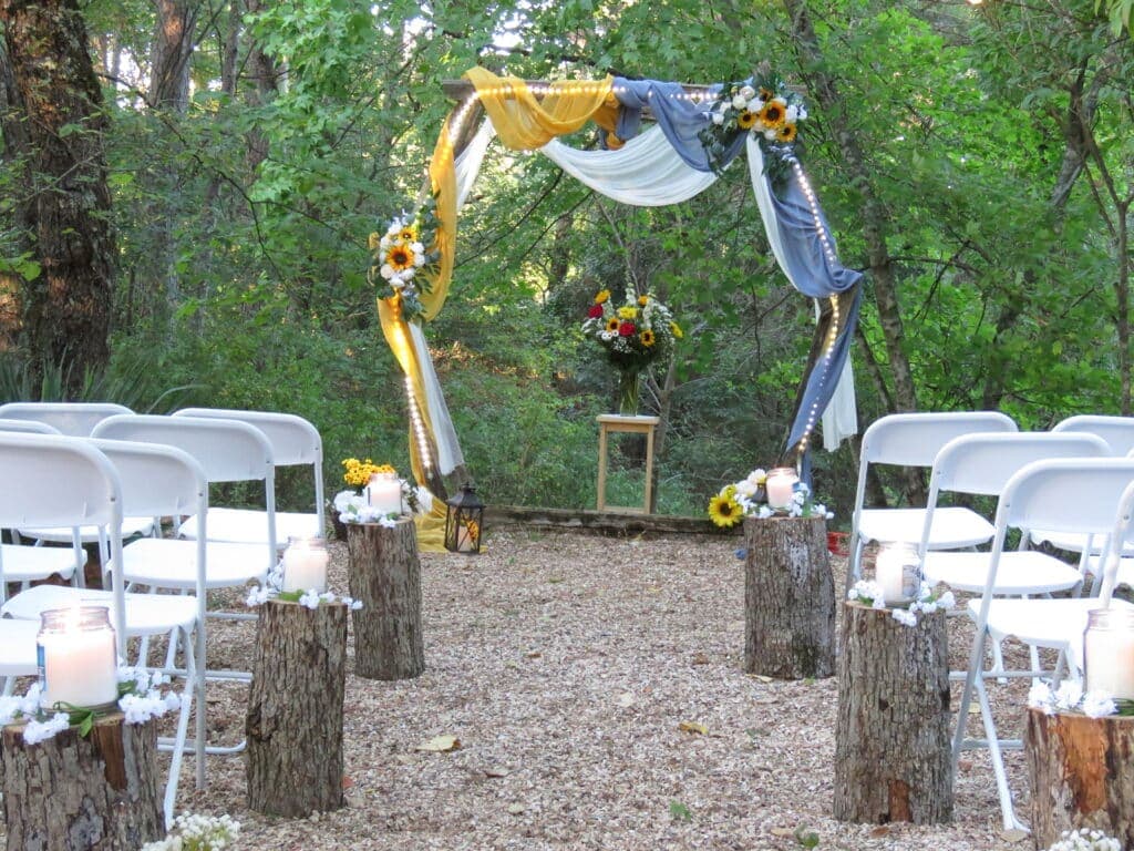 A rustic outdoor wedding ceremony setup with white chairs, a floral arch, and candlelit tree stumps amidst greenery.