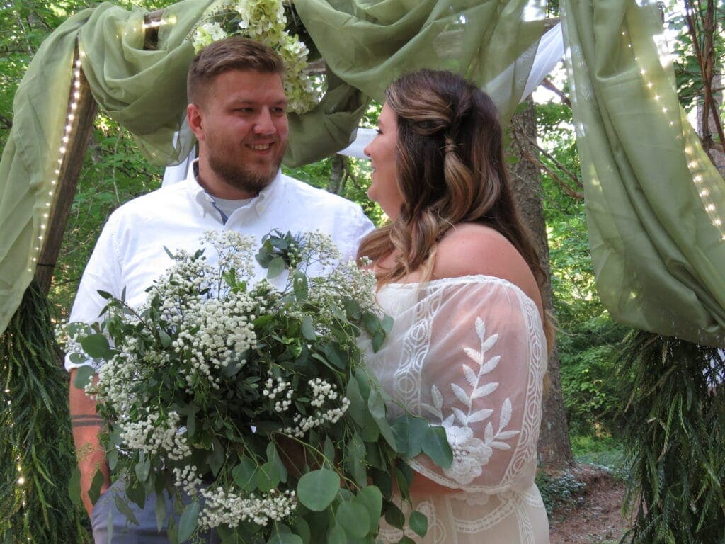 A couple smiles at each other while standing under a green decorative canopy, holding a bouquet of greenery and flowers.