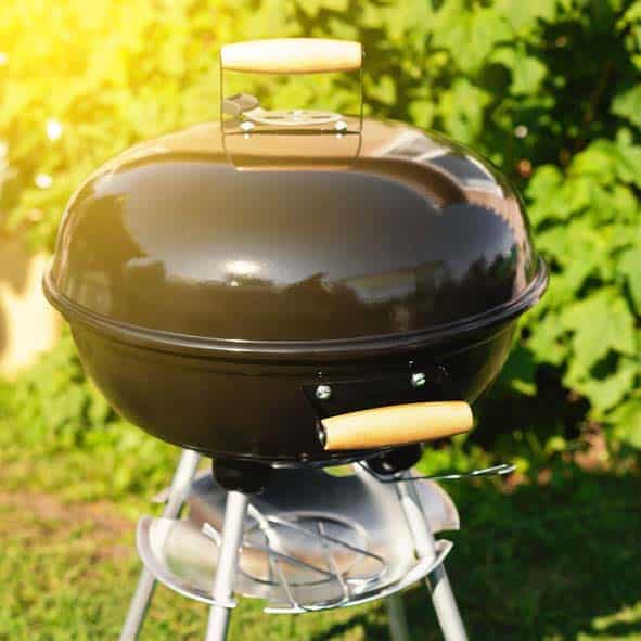 A black charcoal grill sits on a silver stand against a sunny green background.