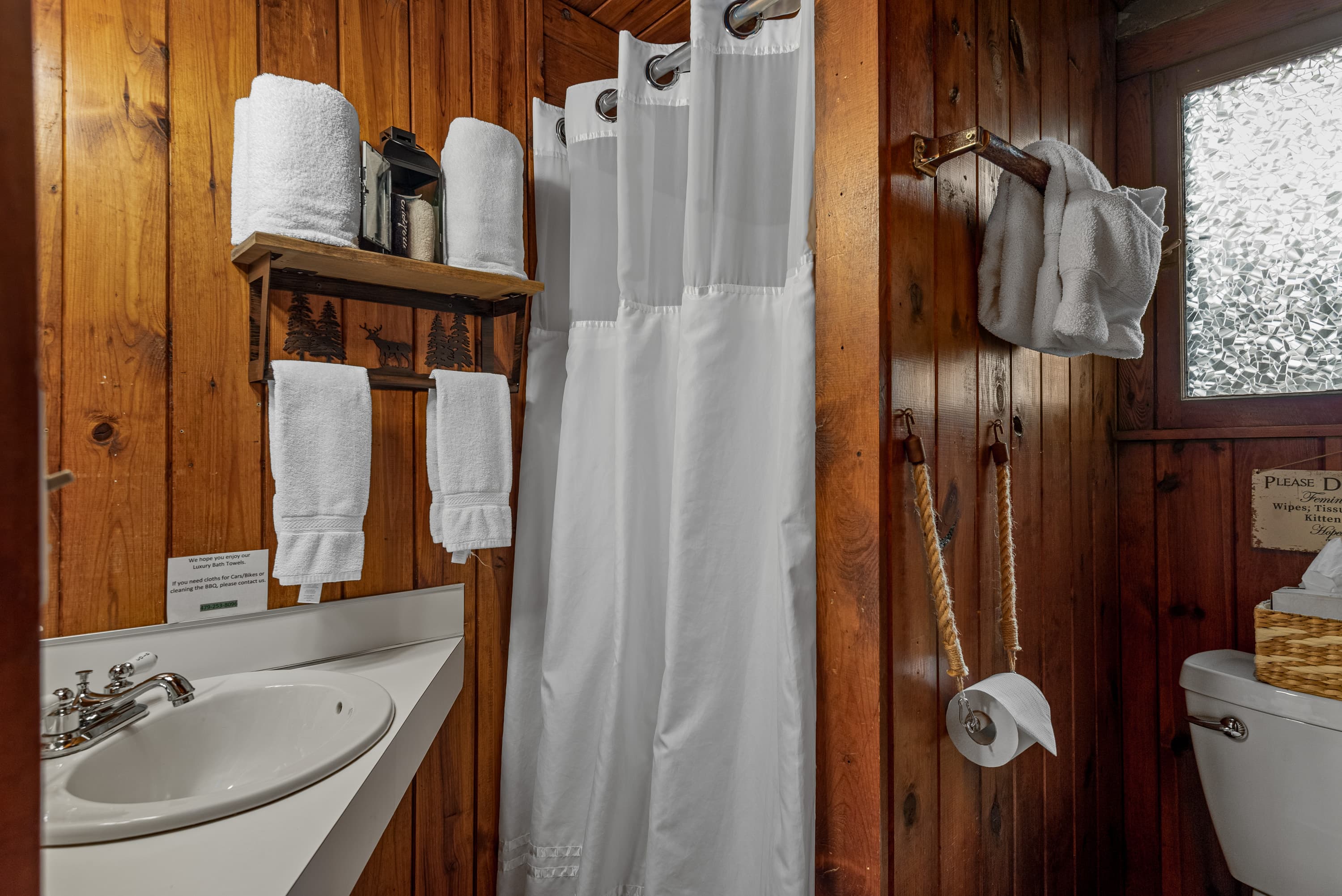 A compact bathroom with wood-plank walls. The space features a sink with a mirror, a toilet, a shower with a white curtain, and a towel rack.