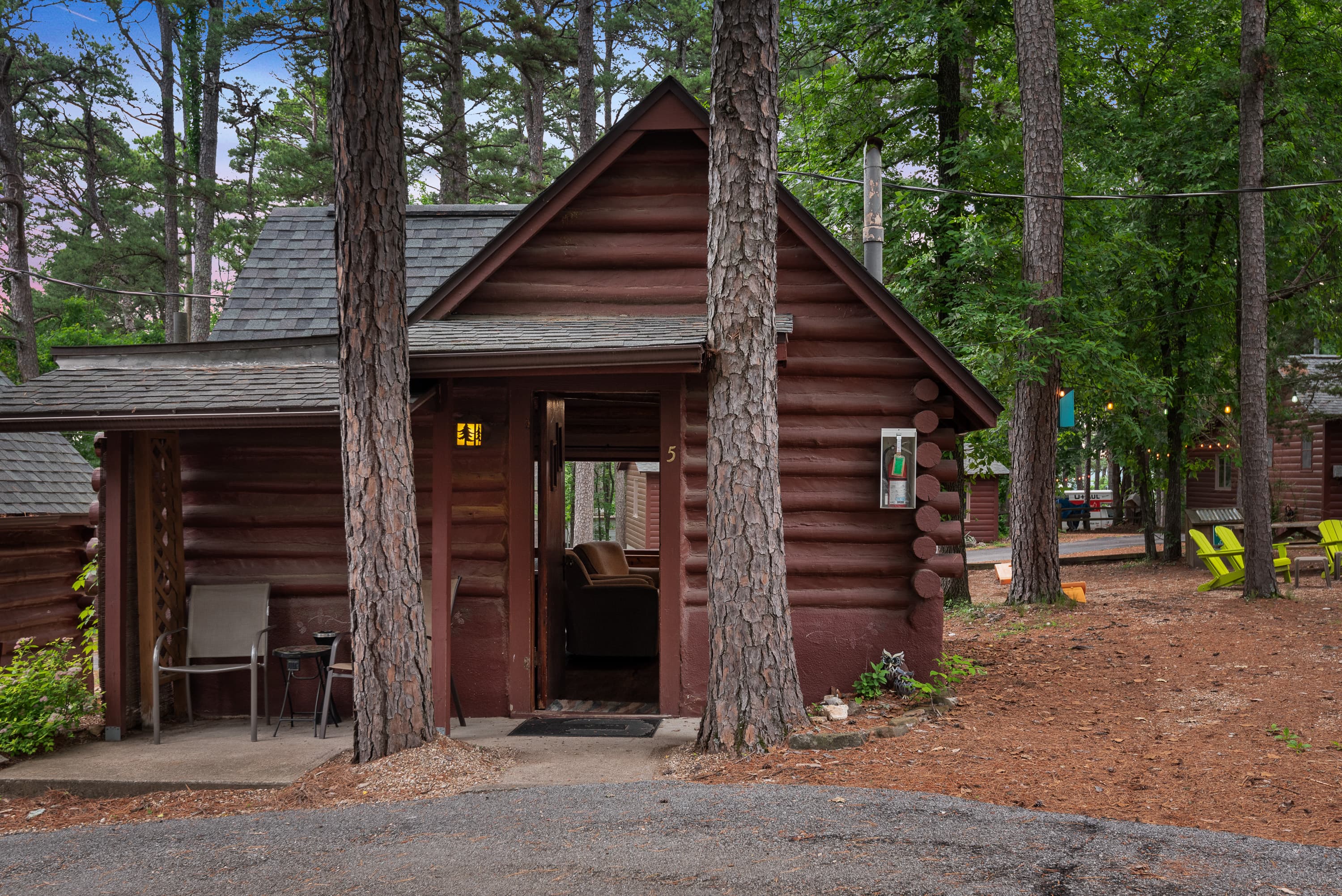 A small, rustic log cabin with a covered porch holding two chairs. The cabin is surrounded by a natural wooded area with pine needles on the ground.