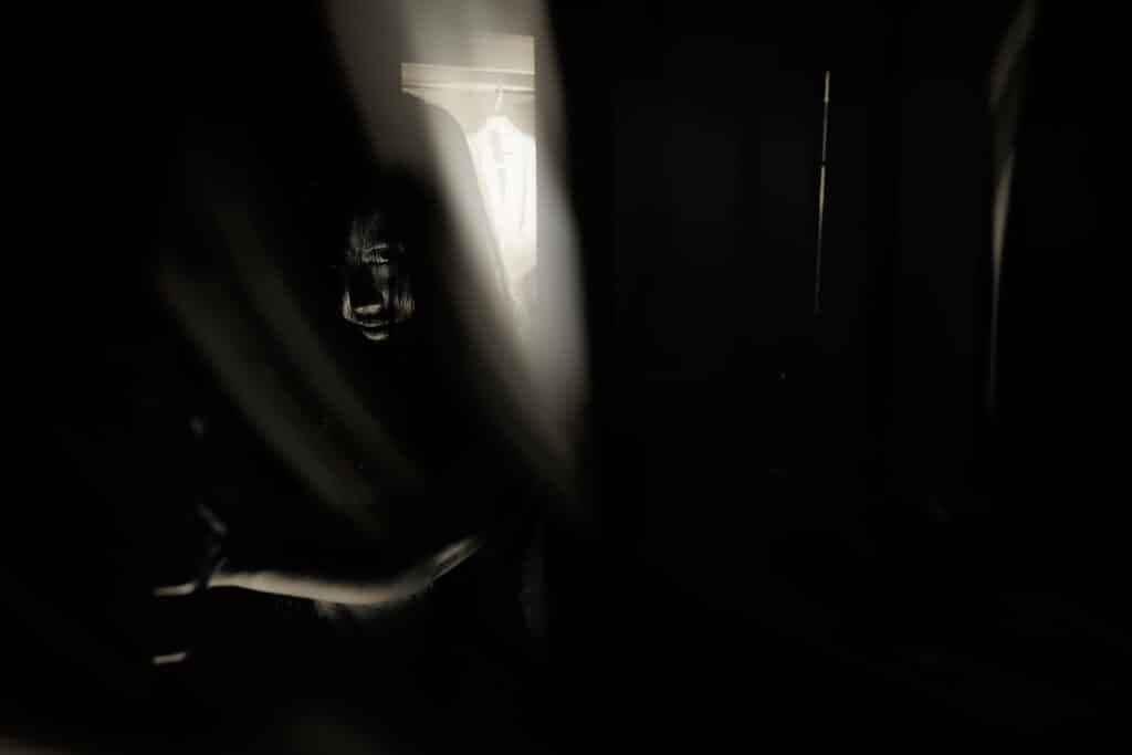 A shadowy figure is partially obscured by curtains, with a faint light illuminating the scene behind them.