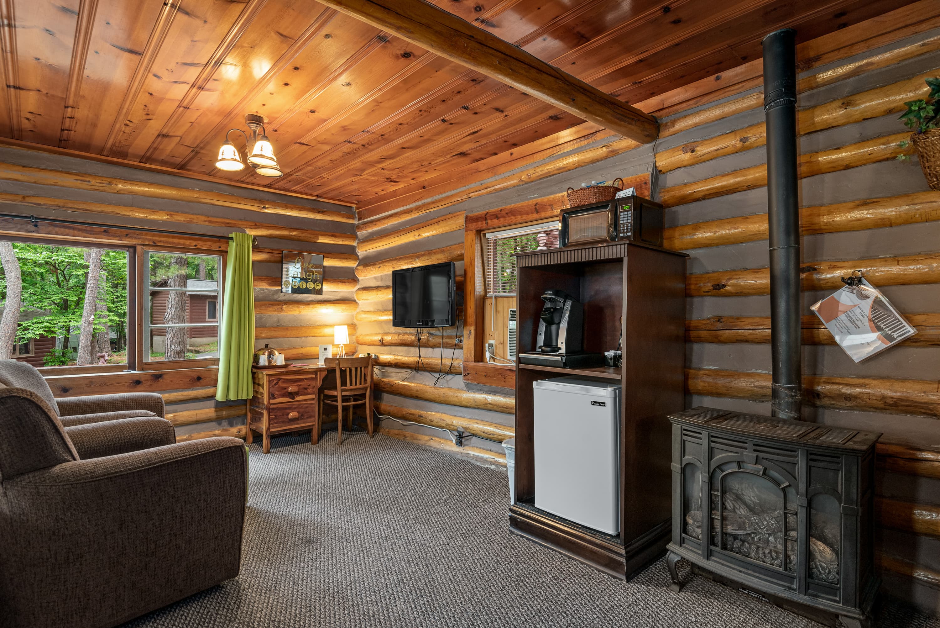 A rustic log cabin room features two armchairs, a television, a small desk, and a gas log stove.