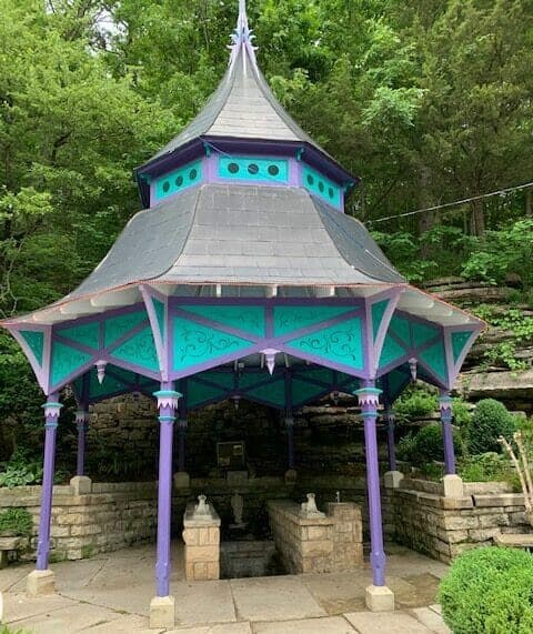 A colorful, ornate gazebo with a pointed roof surrounded by lush greenery.