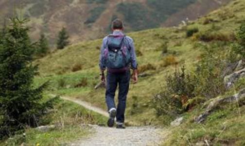 A hiker walks down a winding path in a green, mountainous landscape.