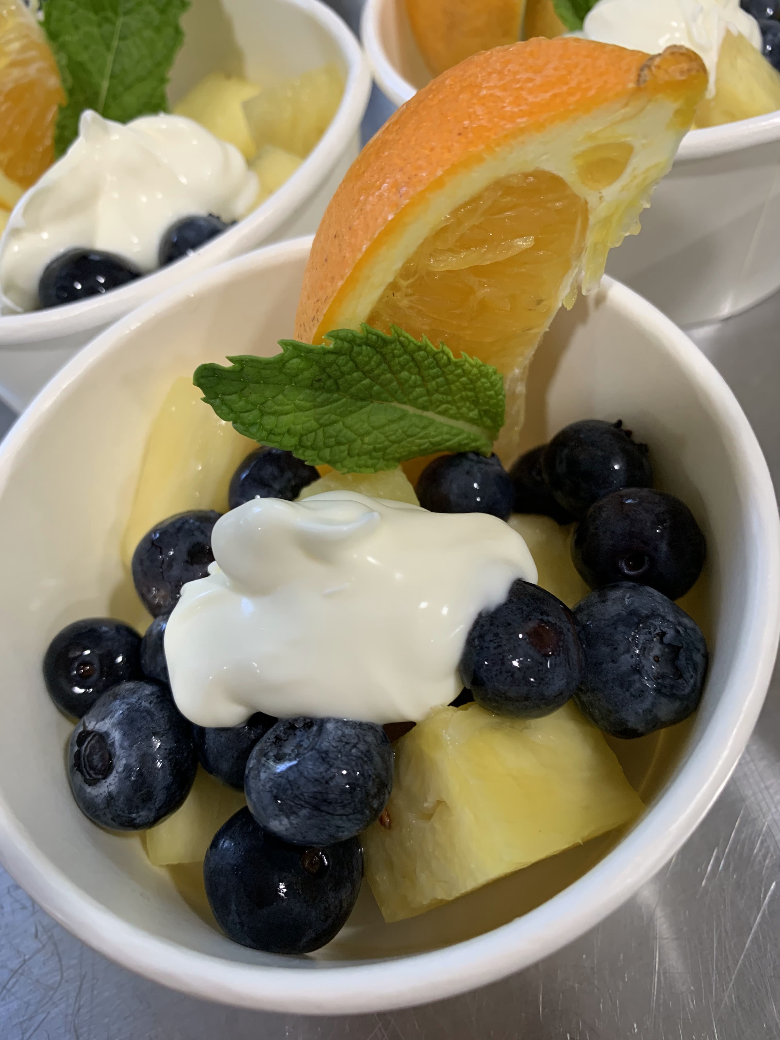A cup of mixed fruit featuring pineapple, blueberries, a slice of orange, and a dollop of cream.