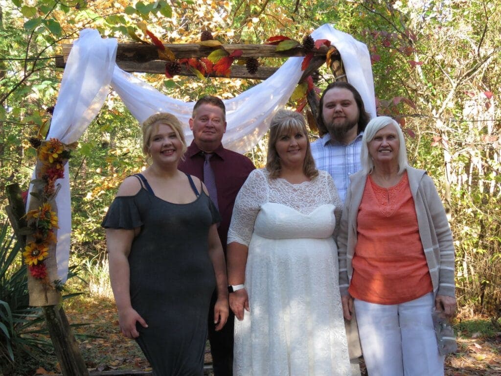 A group of five people, including a bride in a white dress, pose together in front of a decorated arch in a forested setting.