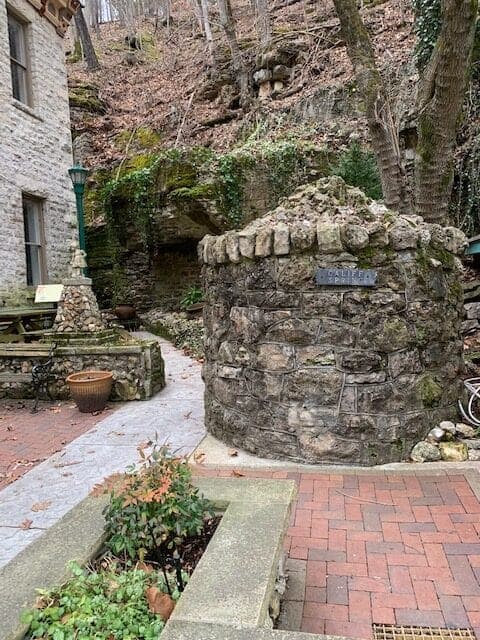 A stone well surrounded by greenery and a brick pathway, near a historic building.