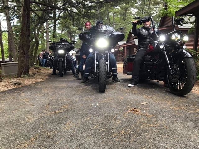 A group of motorcyclists poses with their bikes on a tree-lined path near cabins.