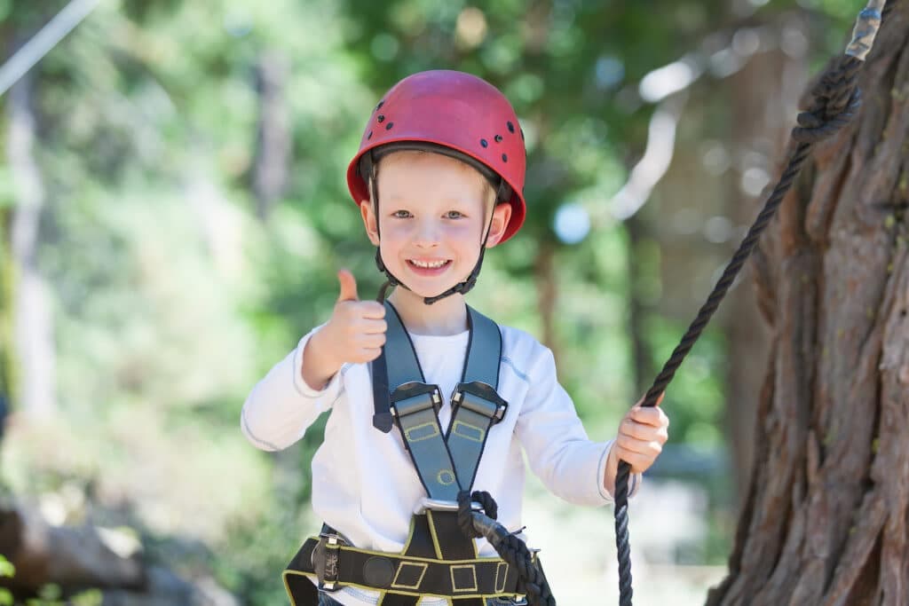 A smiling child in a red helmet gives a thumbs up while holding onto a rope.