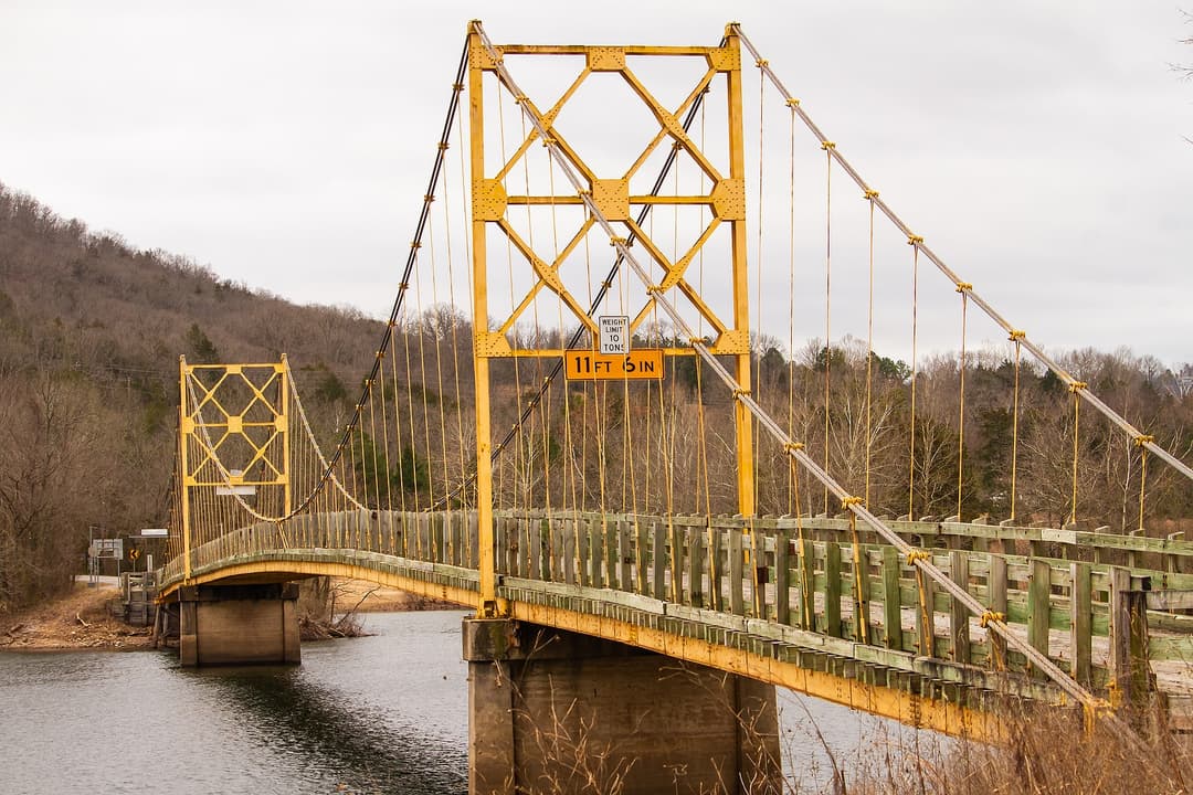 A yellow suspension bridge spans a river with a cloudy sky in the background.