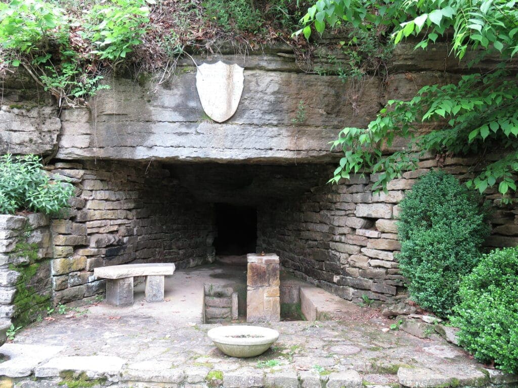 Entrance to a stone cave, framed by greenery and a shield emblem above.