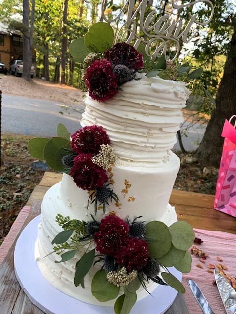 A three-tiered white wedding cake adorned with fresh flowers and greenery, topped with a decorative sign.
