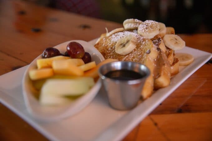 A plate of French toast topped with banana slices and powdered sugar, accompanied by mixed fruit and a small cup of syrup.
