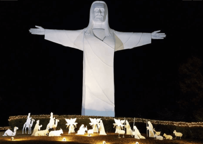 A large white statue of Jesus with outstretched arms stands over illuminated nativity scenes at night.