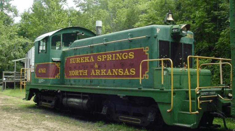 A green train labeled "Eureka Springs & North Arkansas" parked amidst lush greenery.