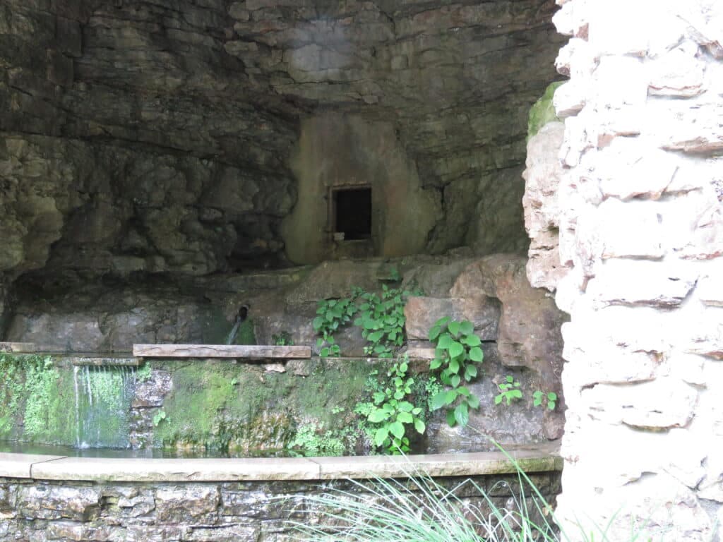 A moss-covered stone cave with a small opening and a water feature surrounded by greenery.