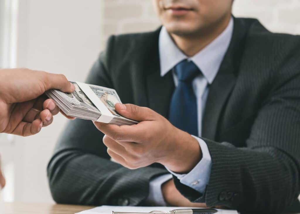 A person hands a bundle of cash to another individual in a suit at a desk.