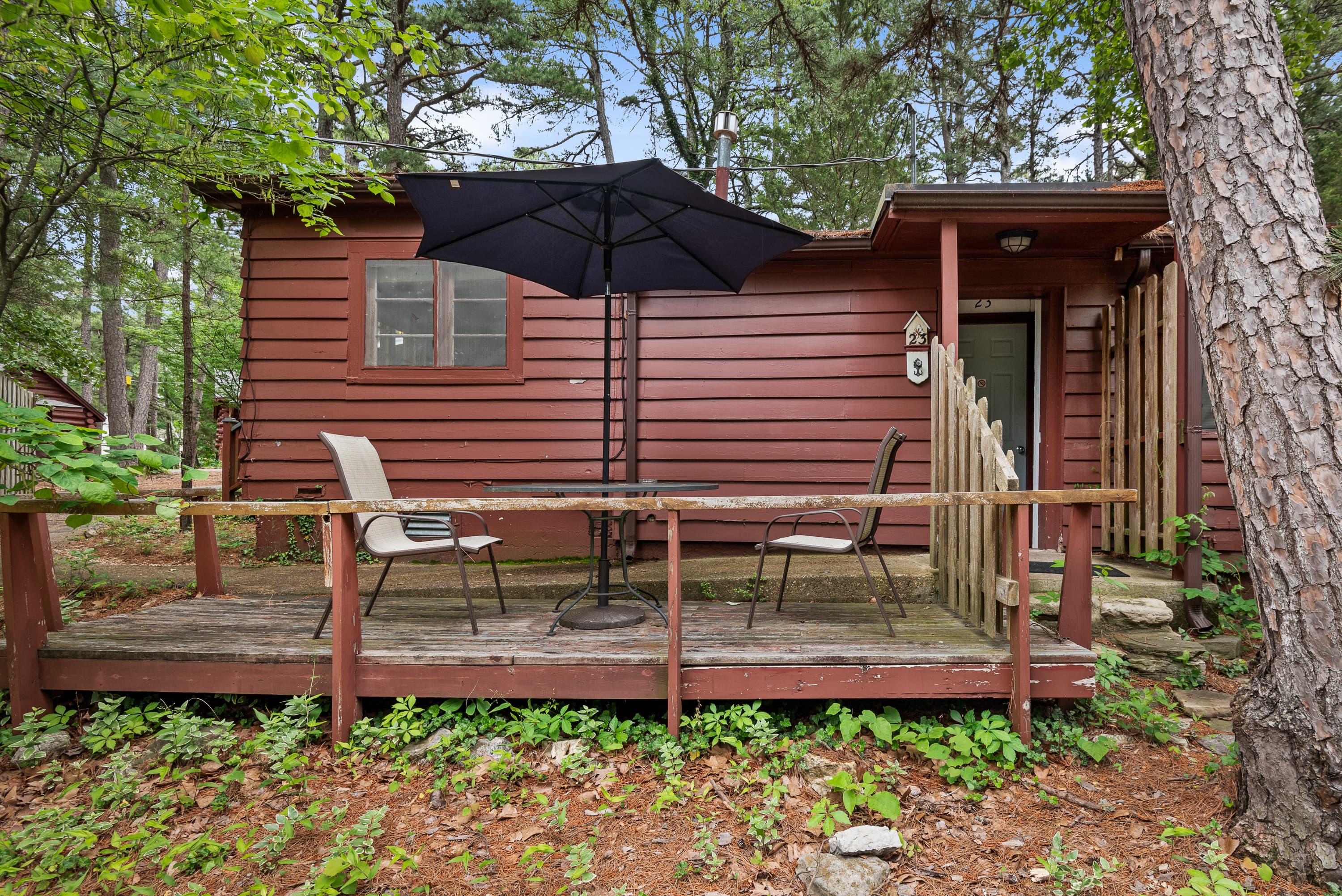 A cozy, rustic cabin with a wooden deck and a black table umbrella. The deck has a small table and two chairs.