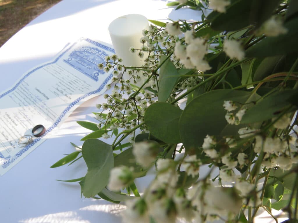 A wedding certificate partially visible among greenery and flowers with a candle in the background.