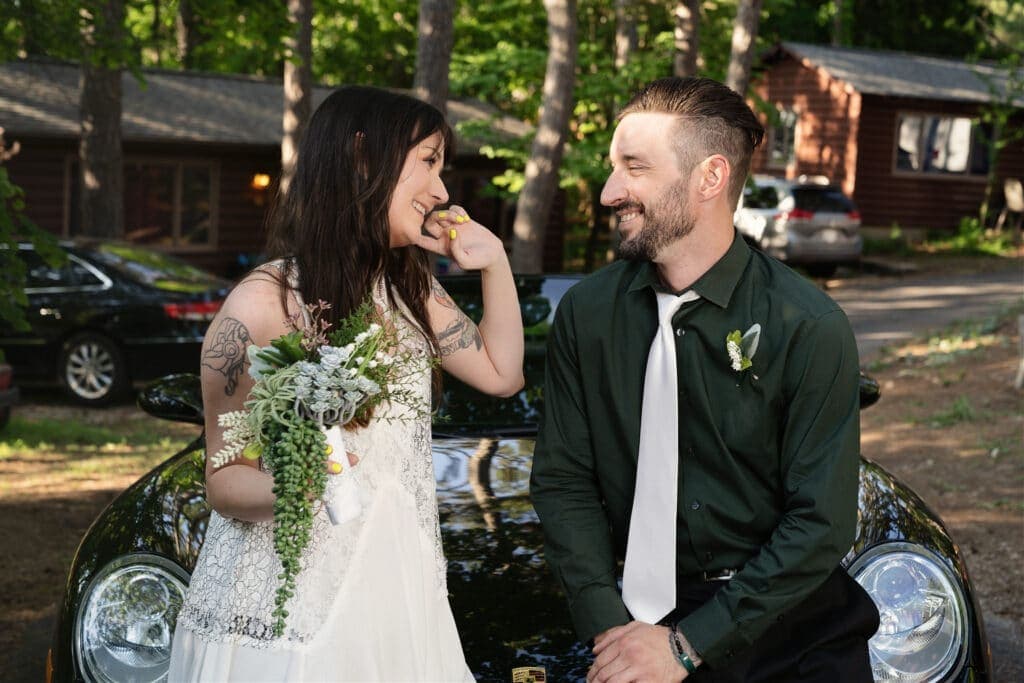 A smiling couple in wedding attire sits on a car, surrounded by a natural wooded setting.