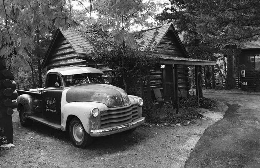 A vintage truck parked outside a rustic log cabin surrounded by trees.