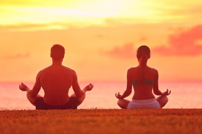 A man and woman meditate together on a grassy shore at sunset.