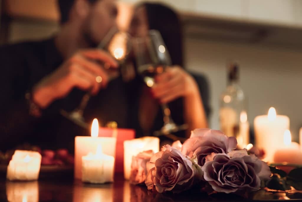 A couple toasts with wine glasses surrounded by candles and roses in a romantic setting.