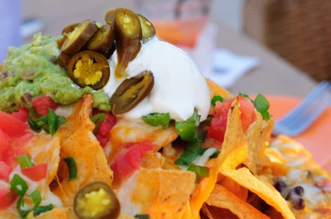 A colorful plate of nachos topped with guacamole, sour cream, jalapeños, tomatoes, and green onions.