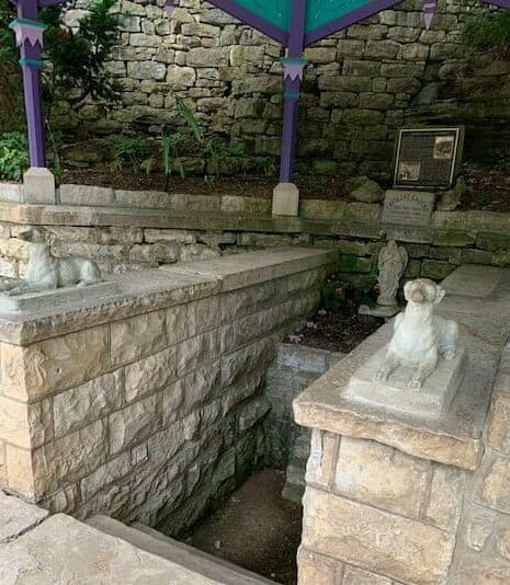 Stone steps leading down to a small grotto, flanked by dog sculptures and surrounded by greenery.
