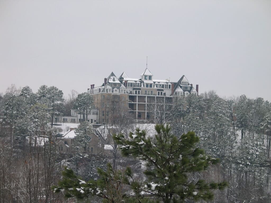 A large historic mansion covered in snow, surrounded by evergreen trees in a winter landscape.