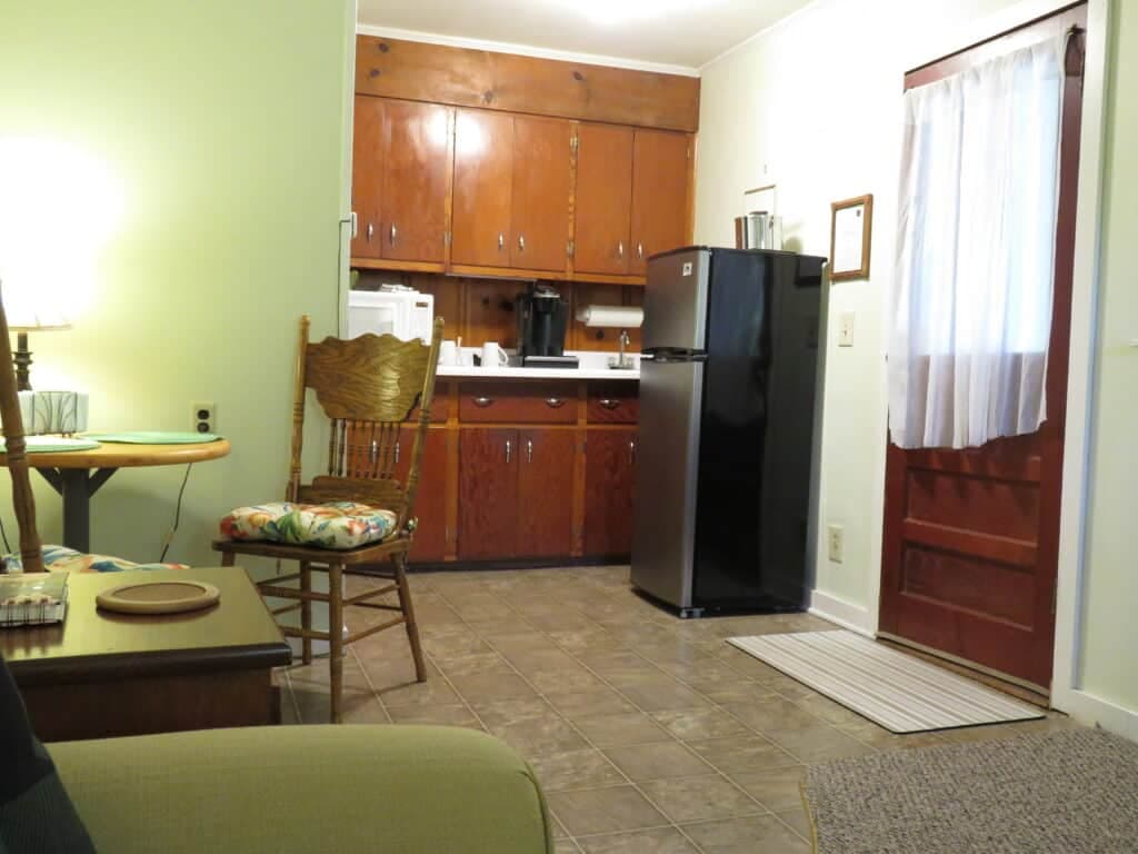 A cozy kitchen and living area featuring wooden cabinets, a black refrigerator, and a comfortable chair.