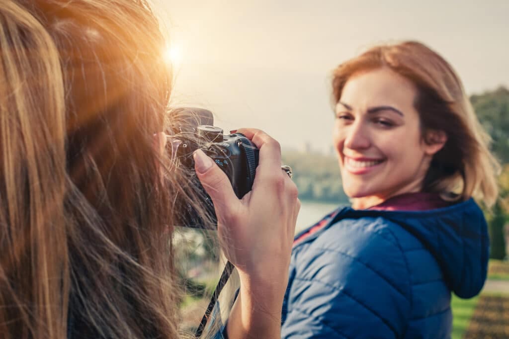 A person takes a photo of a smiling woman outdoors with sunlight in the background.