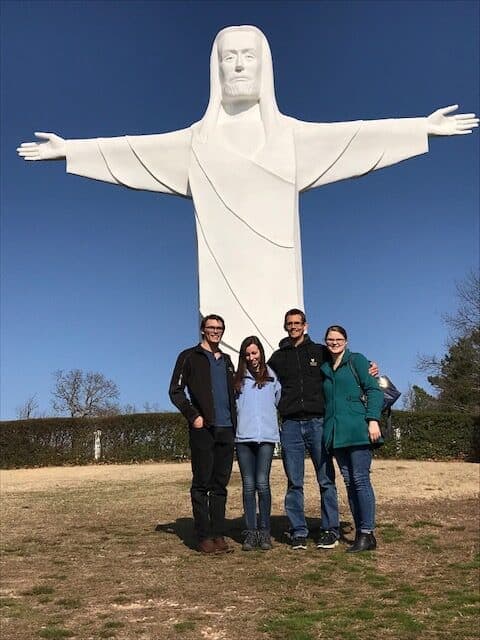 Four people pose in front of a large white statue of Christ with outstretched arms.