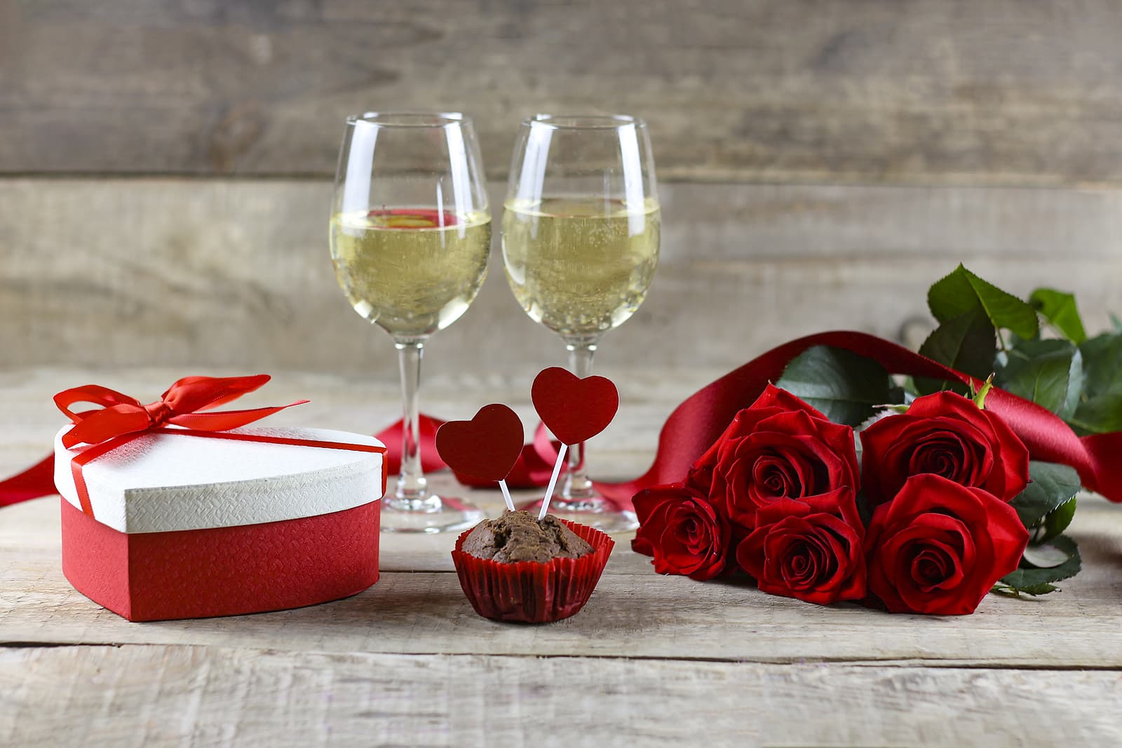 Two glasses of white wine sitting by a heart box of candy and a bouquet of roses